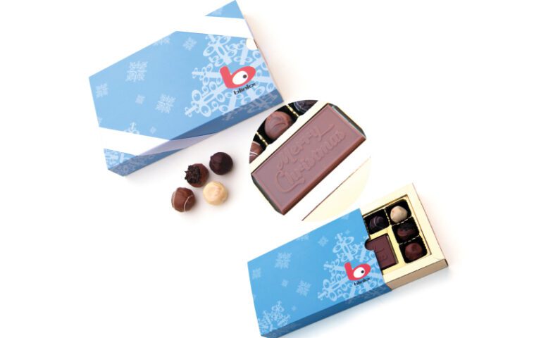 A box of assorted chocolates is partially open, revealing a "Merry Christmas" chocolate bar. The blue box features snowflake designs and a red logo. Three chocolates are outside the box.