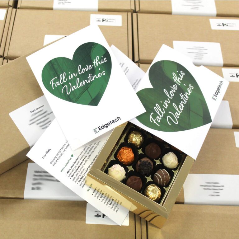 An open box of assorted chocolates sits on top of Valentine-themed cards with "Fall in love this Valentine's" text, surrounded by closed brown packages.