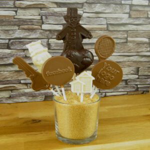 A glass cup holds assorted chocolate lollipops on sticks with different designs, including a gingerbread man, Santa, a guitar, and customizable logos, set against a stone-patterned backdrop.
