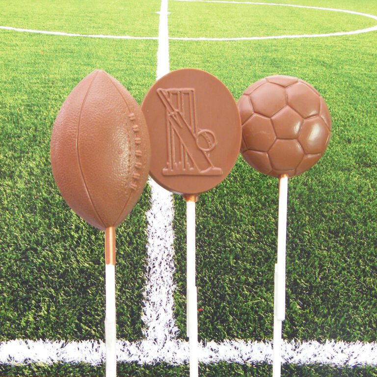 Three chocolate lollipops shaped like a rugby ball, cricket equipment, and a soccer ball, with a grass field background.