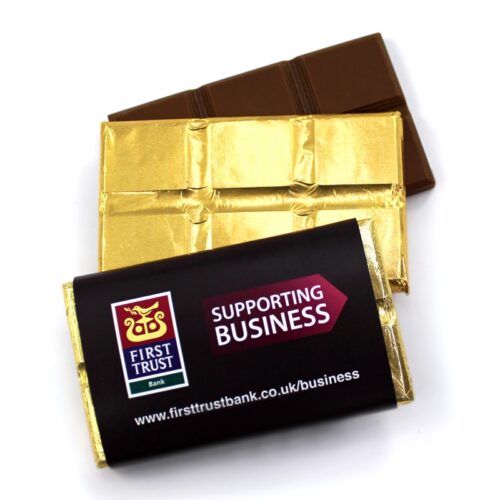 8a_small_break_off_foil_1_7 Two partially unwrapped Small Bar 90 x 50mms chocolate bars, covered in gold foil, display a "First Trust Bank" logo and the text "Supporting Business.