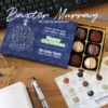 A box of chocolates with a "Happy Christmas" card from Ulster Bank lies open on a wooden desk, next to a pen, keyboard, and printed gift notes.