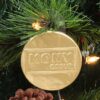 Gold foil coin with "MONY GROUP" embossed on it, hanging on a Christmas tree with lights in the background.