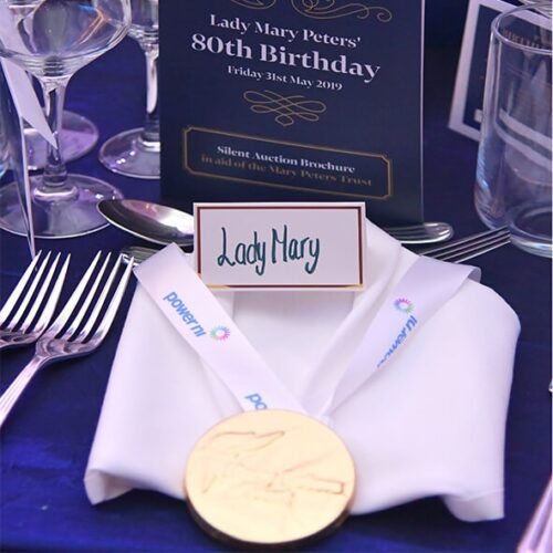 Elegant table setting features a place card reading "Lady Mary," a folded napkin with a Corporate Chocolate Medal on top, and an announcement card for Lady Mary Peters' 80th Birthday on May 31, 2019.