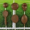 img_1421_optimized Five chocolate lollipops shaped like sports items: golf ball, soccer ball, boxing glove, tennis racket, and football, displayed on a grassy background.