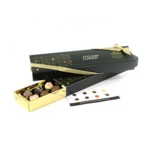 The Sixteen Chocolate Customised Sleeve Box is open, showcasing a variety of chocolates. It features a long, rectangular design in black and gold with a decorative gold ribbon and an accompanying description card.