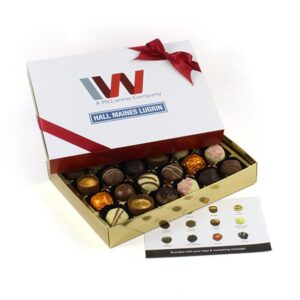 A partially open Twenty Four Chocolate Customised Box Sleeve displays assorted truffles, adorned with a red ribbon and company logos on the lid. A card showcasing chocolate images sits beside it.
