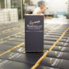 A box labeled "Gold Hot Foil Printing" sits on a production line with similar boxes, in a factory setting with a worker in the background.