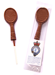 A chocolate lolly shaped like a tennis racquet next to its clear packaging, which displays a royal-themed label with a crown and text.