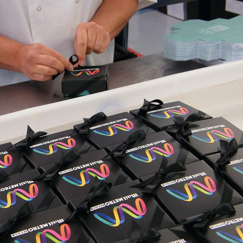 A person is tying a black ribbon on a small black gift box. Several similar boxes are arranged neatly on a table, each with a colourful Radio Metro 105.9 FM logo on the lid.