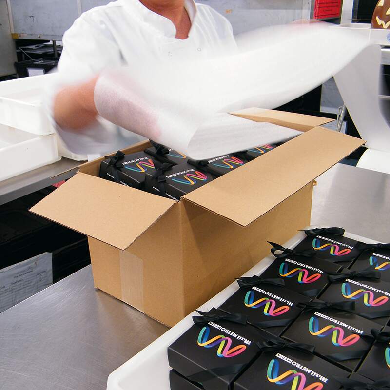 A person in a white coat places black boxes with colourful designs and the text GOOD OLNEWT into a cardboard box on a stainless steel counter. More boxes are stacked nearby.