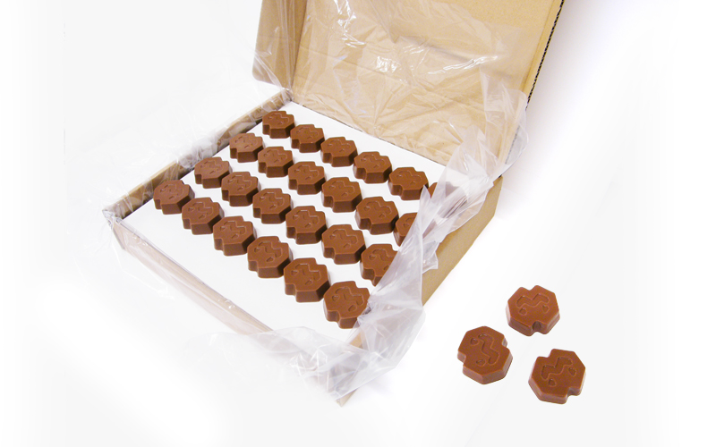 A cardboard box lined with plastic holds rows of octagonal, brown chocolates with geometric patterns. Three chocolates are placed outside the box on the right side.