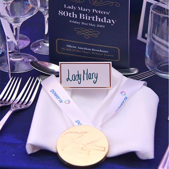 A CORPORATE EVENT DINNER TABLE PLACE SETTING WITH CUTLERY, GLASSES AND A WHITE LINEN NAPKIN FOLDED TO RESEMBLE CLOTHING, ONTO OF THIS is a bespoke logo chocolate medal with a branded ribbon, showing the role of chocolate in event branding.