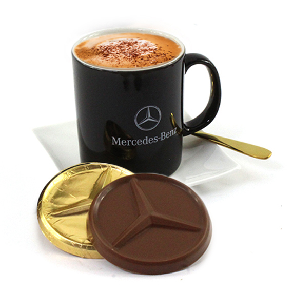 A Merceedes branded black coffee cup on a white saucer with a gold spoon on the side and leaning on the saucer is a large chocolate coin with a Mercedes logo, a perfect bespoke marketing gift