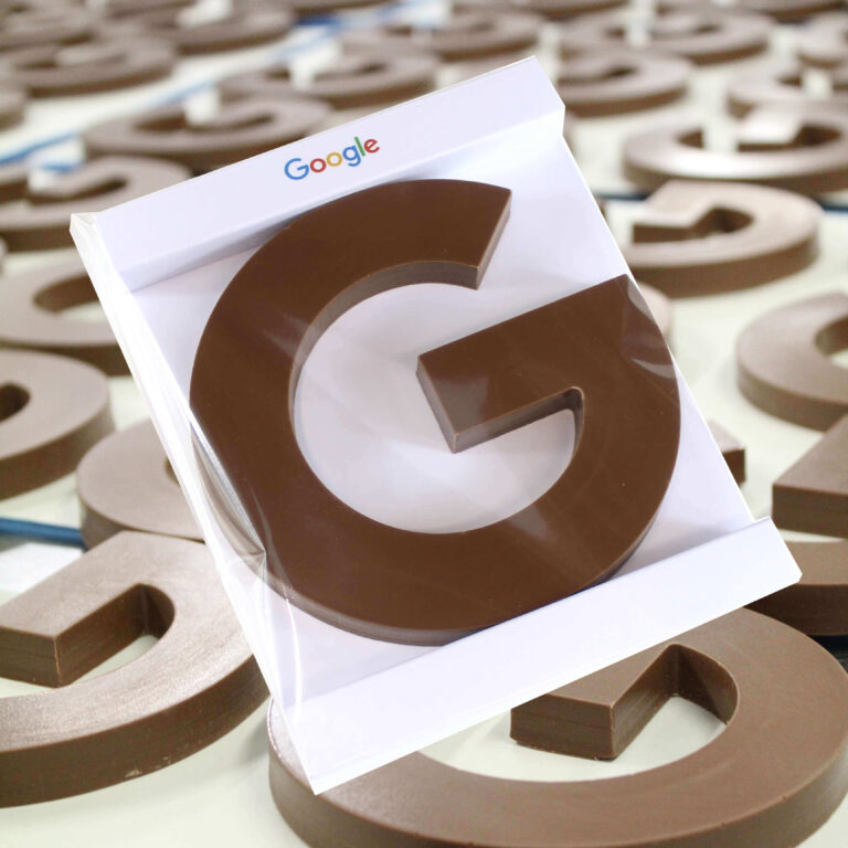 A large chocolate letter G, for Google displayed flat on a table top with a G packaged in Google branded packaging in the centre, a handmade corporate chocolatier gift